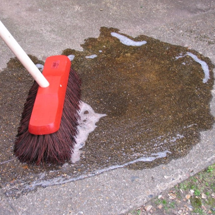 A red push broom with dark brown bristles and a light-colored handle cleaning a wet patch of dark soil or debris on concrete pavement.