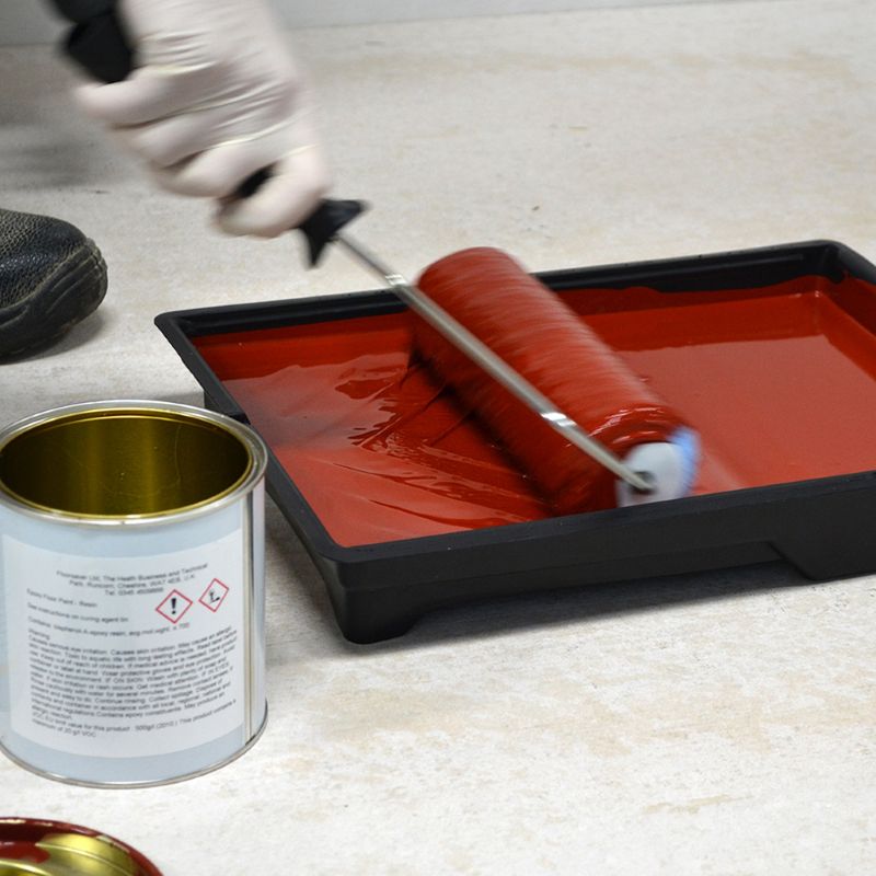 Person painting a surface with red paint using a roller and can of paint on a light-colored floor.